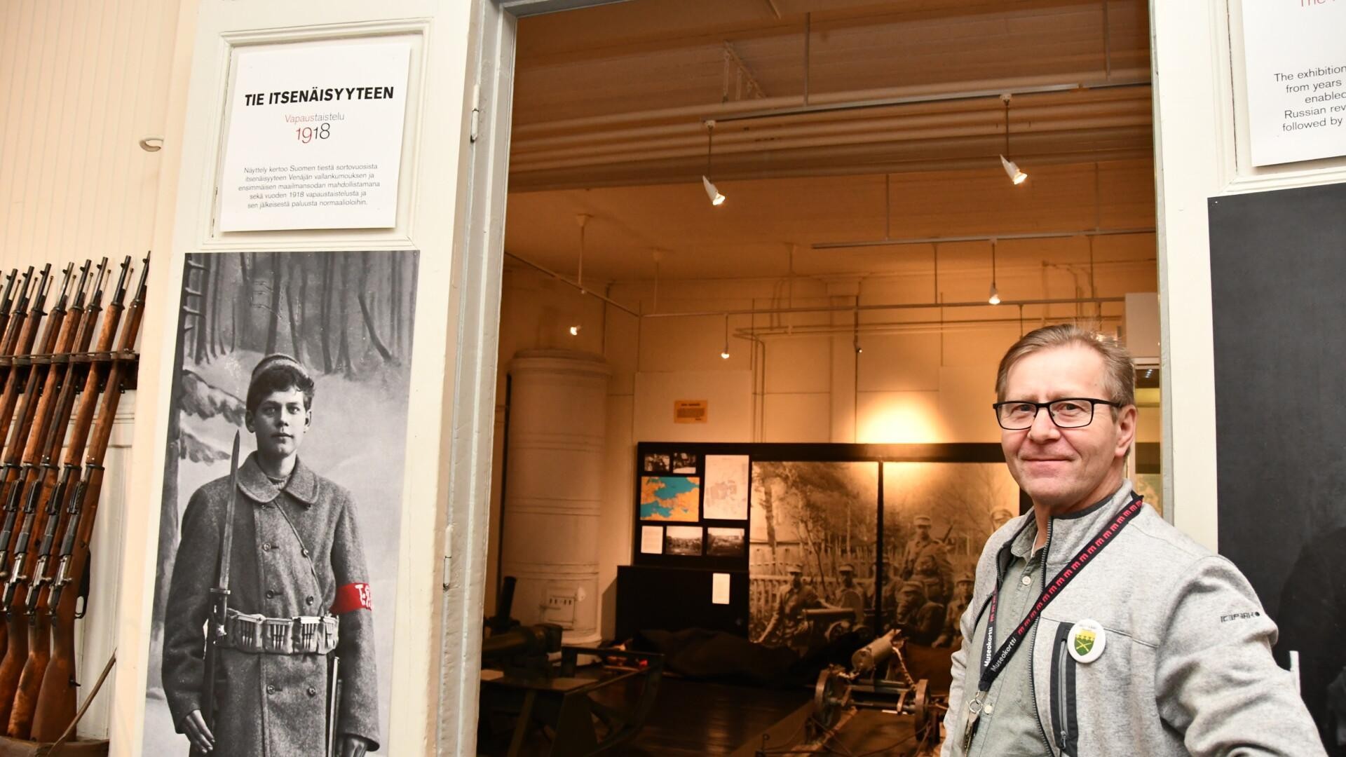 Mies seisoo näyttelyn oviaukolla, vieressään vanha kuva sotilaasta