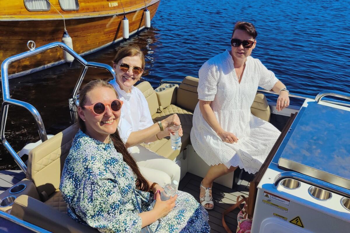 Kolme naista aurinkolasit päässään istuu moottoroidun veneen penkissä, takana näkyy laiturissa pieni perinteinen puulaiva.
