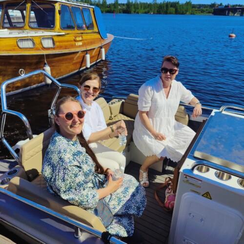 Kolme naista aurinkolasit päässään istuu moottoroidun veneen penkissä, takana näkyy laiturissa pieni perinteinen puulaiva.