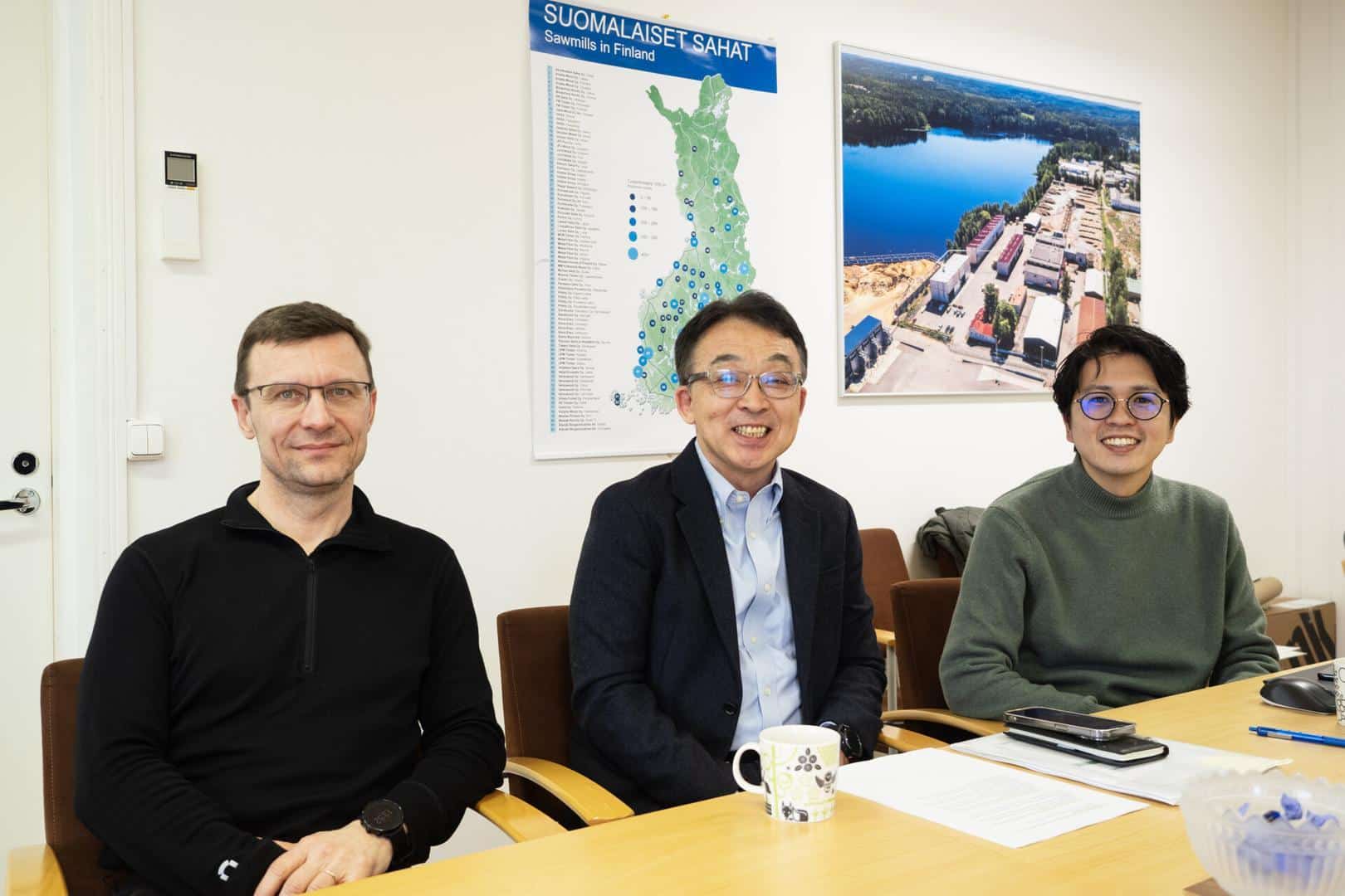 Kolme miestä istuu vierekkäin neuvottelupöydän äärellä, takanaan kaksi taulua