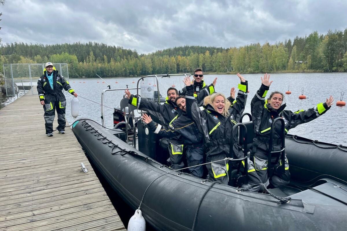 tausta metsää ja järvi, kumiveneessä seisoo joukko kumipukuisia ihmisiä kädet pystyssä