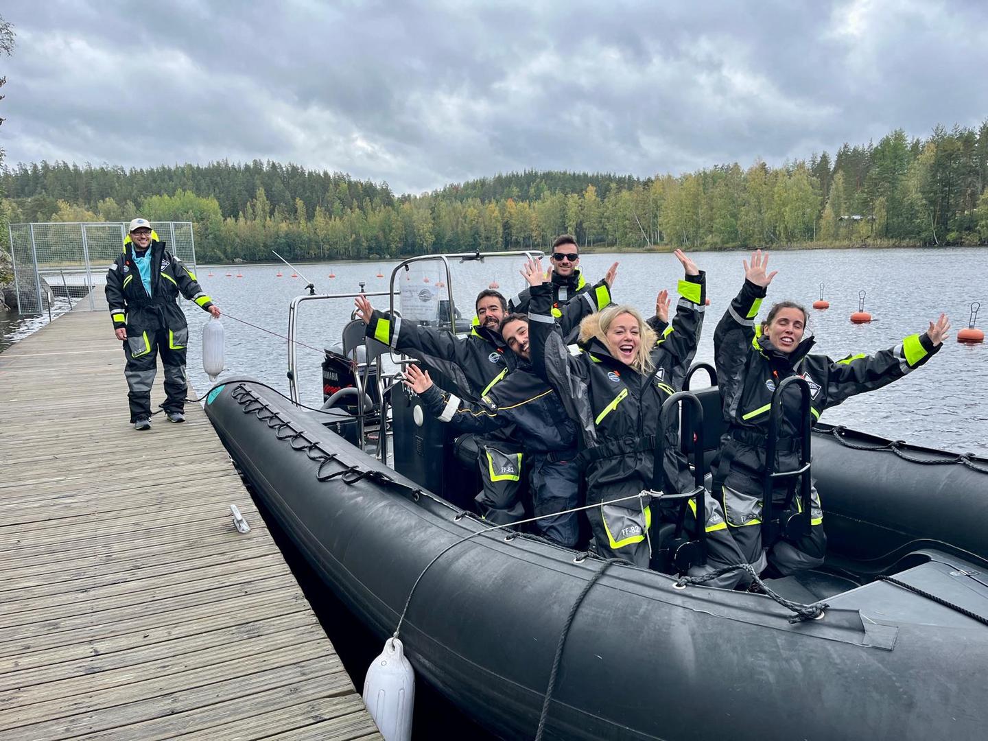 tausta metsää ja järvi, kumiveneessä seisoo joukko kumipukuisia ihmisiä kädet pystyssä