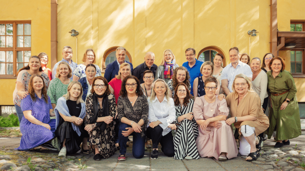 Ihmisiä kolmessa rivissä katsomassa kameraan, taustalla keltainen talonseinä