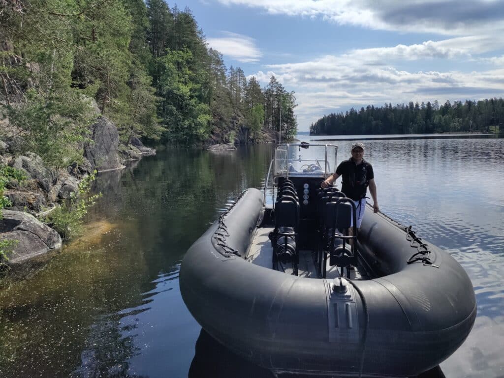 Musta kumine vene, jossa seisoo mies. Taustalla järvi ja metsämaisema.