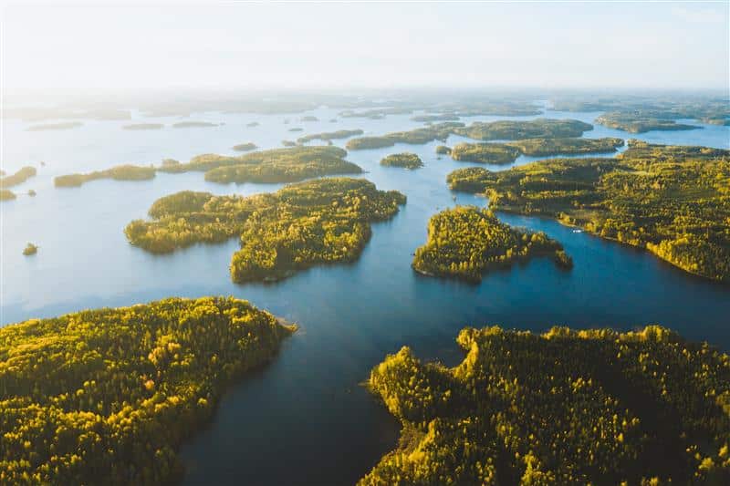 Kuva on ilmakuva Saimaan saaristosta, aurinkoisessa kuvassa näkyy laaja sininen järvimaisema ja pieniä vehreitä saaria.